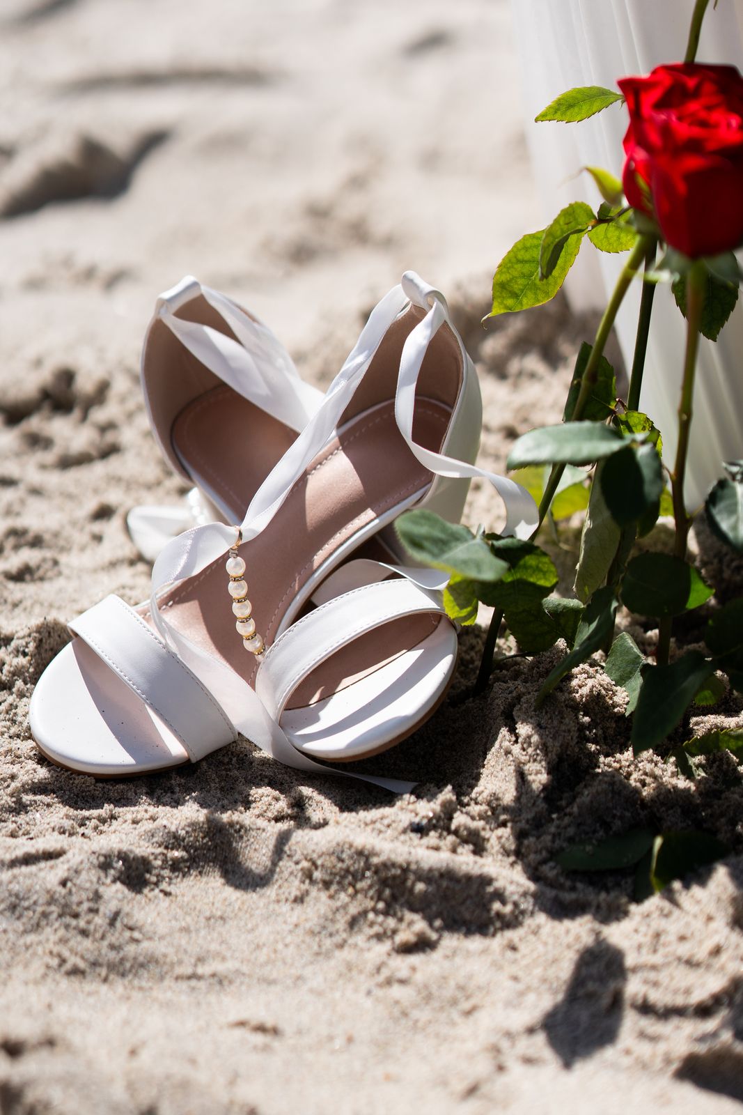 White bridal sandals in the sand beside a red rose