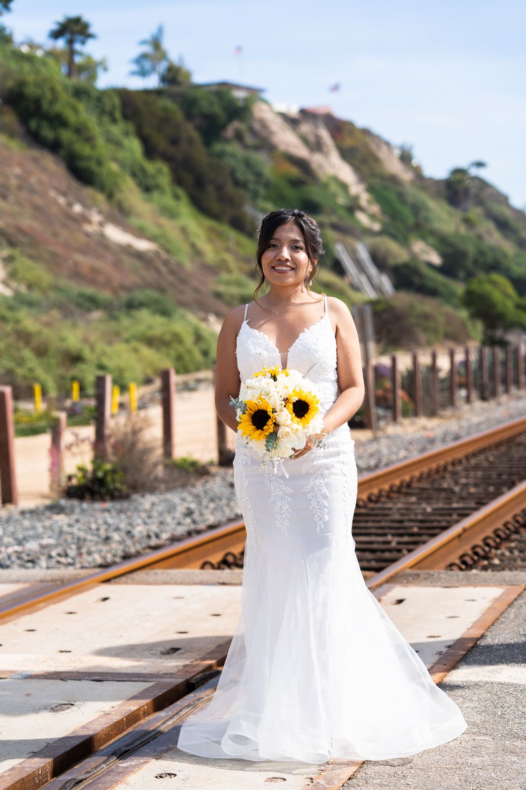 Bride holding a sunflower bouquet by coastal railroad tracks