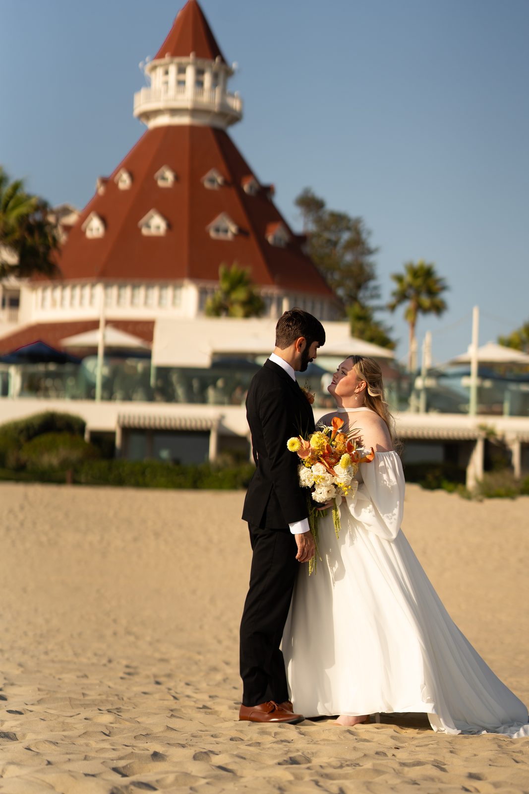 Bride and groom on the beach in front of a historic red-roofed hotel