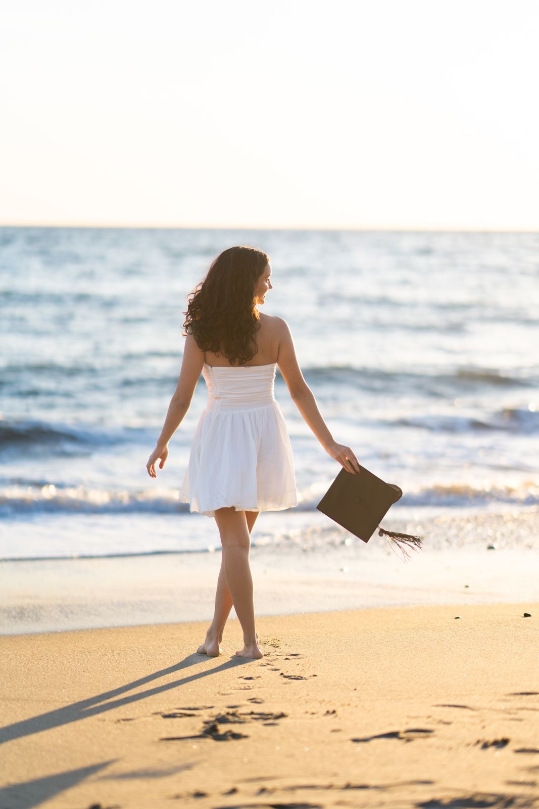 Graduate in a white dress walking barefoot along the shore holding her cap