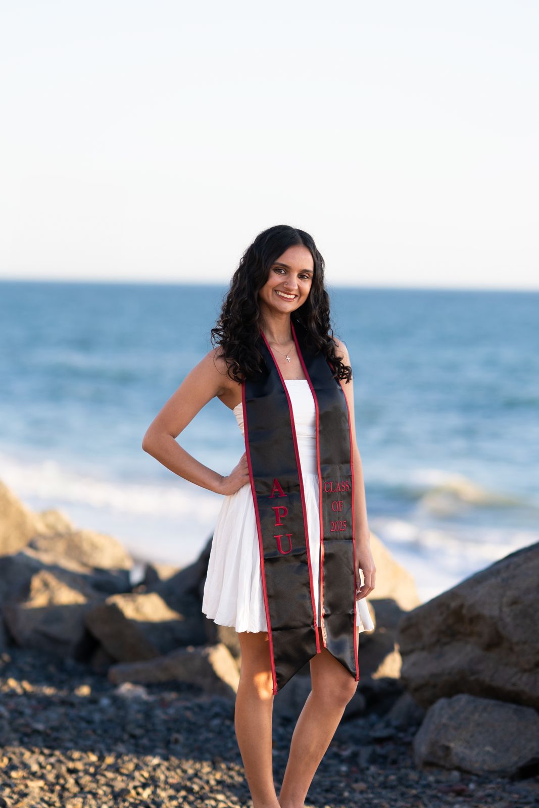 Smiling graduate in an APU Class of 2025 stole by the ocean